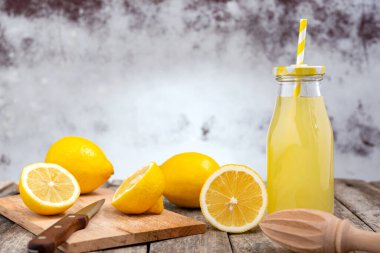 Bottle of cold lemonade with drink straw and cutting board with fresh lemon cut in half and knife on a rustic wooden table. Selective focus.
