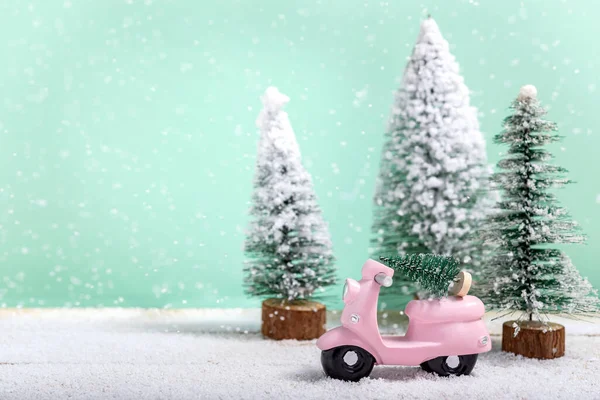 Pink toy motorbike with a Christmas tree next to fir trees while it is snowing on a wooden table. Concept of celebrating Christmas and  New Year.