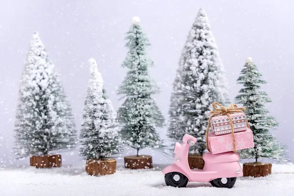 Pink toy motorbike with a Christmas gifts next to fir trees while it is snowing on a wooden table. Concept of celebrating Christmas and New Year.
