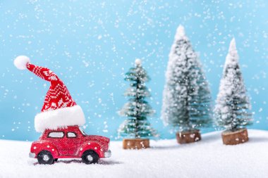 Kar yağarken çatıda Noel Baba şapkalı kırmızı oyuncak araba köknar ağaçlarının yanında. Kış ve Noel konsepti.