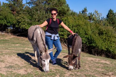 Gülümseyen bir turist kadın otlakta otlayan eşeklerin yanında poz verir..
