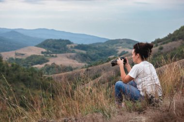 DSR kameralı kadın fotoğrafçı doğanın güzel manzaralı bir tepedeki uzun çimlerde oturuyor. Seyahat ve hobi kavramı.