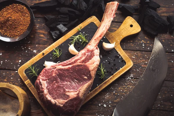Dry aged raw tomahawk black angus prime beef chop with salt, pepper, rosemary, garlic, charcoal and a knife