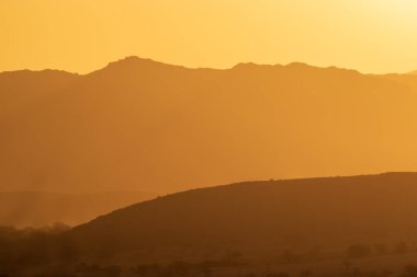 Sıcak bir günbatımı uzak dağların siluetlerini aydınlatır. Altın renkler huzurlu ve huzurlu bir çöl atmosferi yaratır.