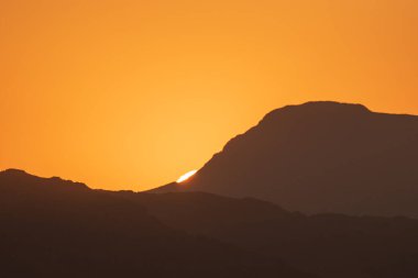 Sıcak bir günbatımı uzak dağların siluetlerini aydınlatır. Altın renkler huzurlu ve huzurlu bir çöl atmosferi yaratır.