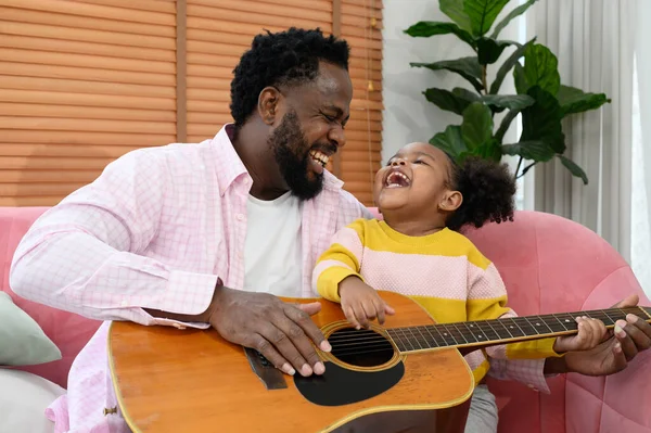 African girl and father are playing guitar while sitting on the sofa at home