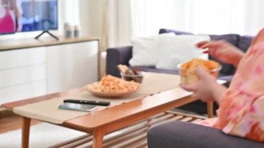 4K. Şişman kadının elinde patates cipsiyle kanepede televizyon izlerken yakın plan.