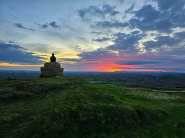 Hartshead Pike, Ashton-under-Lyne 'de canlı bir günbatımını izleyen yalnız bir figürün sakin bir fotoğrafı. Sahnede bir taş anıt, yeşil tepeler ve renkli bir gökyüzünün altında uzak bir şehir silueti yer alıyor. Yalnızlık ve doğa temaları için mükemmel.