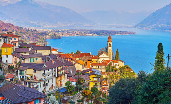The old tile roofs, colored houses and tall bell tower of San Martino Church in Ronco sopra Ascona against Lake Maggiore and Alps in haze, Switzerland