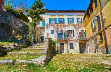 The small shabby Piazza Rusciett square with broken stone stairs, medieval townhouses and grass overgrown through the cobblestone, Castello, Valsolda, Italy