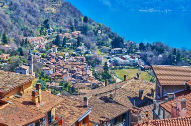 The small Loggio settlement with belfry of San Bartolomeo Church and small houses behind the roofs and chimneys of Castello, Valsolda, Italy