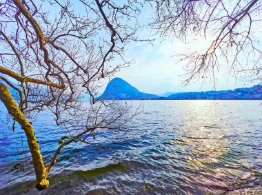 Lugano Gölü 'nün dalgalı yüzeyi ve Monte San Salvatore' un silueti altın alacakaranlıktaki çınar dallarının arkasında, Lugano, İsviçre