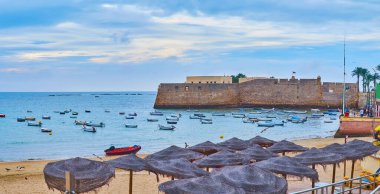 Cadiz Körfezi Panoraması La Caleta plajında saman güneş gözlüğü ve arka planda Santa Catalina Kalesi 'nin taş kaleleri, Cadiz, İspanya