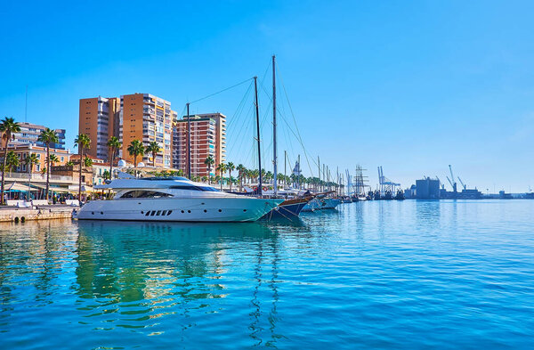Ярко-голубые сияющие воды порта Малага, отражающие яхты и здания на пирсе Мюэль Уно, Испания