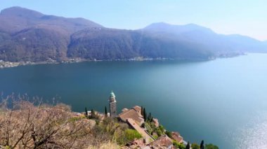 The slender stone bell tower of Santa Maria del Sasso Church against azure Lake Lugano and Alpine scenery, Morcote, Switzerland