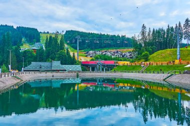 Aynaya benzeyen Trout Pond 'un etrafındaki yeşil dağ yamaçlarında restoranlar ve turistik oteller, Bukovel, Karpatlar, Ukrayna