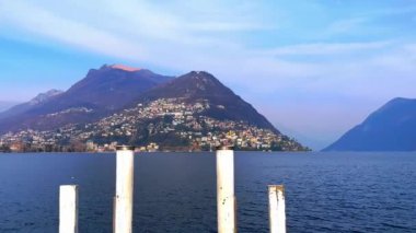 Lugano Gölü 'nün rıhtımı gölün parlak mavi dalgalı yüzeyi, Monte Bre, Monte Boglia ve arka planda puslu Alpler, Lugano, Ticino, İsviçre