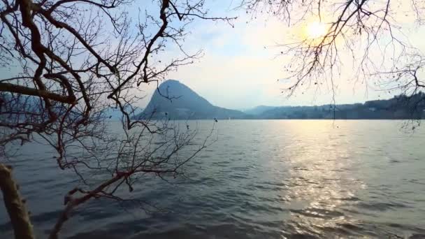 ルガーノ スイス ルガーノ バックグラウンドでモンテサルヴァトーレとシカモアの枝を通して熟したルガーノ湖の美しい夕日ビュー — ストック動画