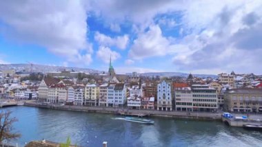 Lindenhof Tepesi 'nden Zürih silueti üzerinden hızla akan bulutların zamanlaması, feribot, limatquai seti, konak evleri, karlı çatılar ve arka planda Niederdorf, İsviçre' deki Predigerkirche Kilisesi 'nin kulesi ile Limmat Nehri' ni gözlemliyor.