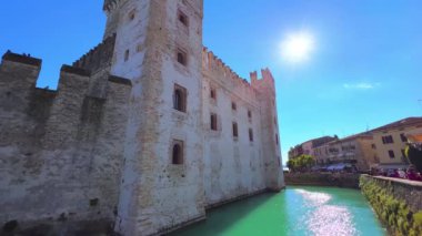 The sunny sky over the medieval Scaligero Caslte and the dug moat around it, reflecting the sun path on its rippled waters, Sirmione, Italy