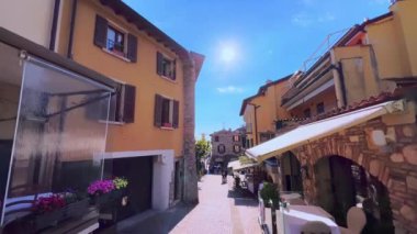 Walk down narrow old Via San Salvatore street with outdoor cafes, restaurants and bars, decorated with flowers in pots and green plants, Sirmione, Italy