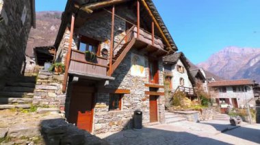 Kilisenin saat kulesi, taş evleri ve dağları olan güneşli caddenin panoraması, Sonogno köyü, Valle Verzasca, İsviçre