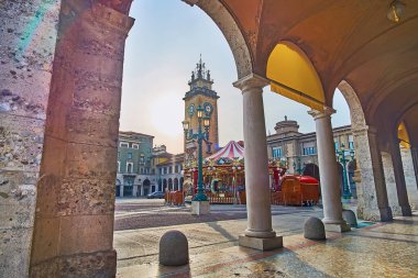 The tall stone Memorial tower and vintage carousel from the portico of Piacentiniano Centre, Piazza Vittorio Veneto, Citta Bassa, Bergamo, Italy clipart