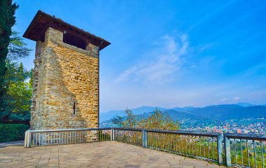Rocca di Bergamo kalesinin terası Sant 'Eufemia tepesinin tepesinde taş gözetleme kulesi manzaralı ve arka planda puslu Bargamasque Alpleri, İtalya