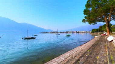 Maggiore Gölü etrafında puslu dağlar, dalgalı sularda sallanan küçük kayıklar ve yatlar ve göldeki çam ağaçları, Locarno, İsviçre