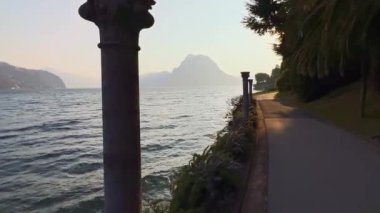 Resimli Park Villa Heleneum 'da Ceresio göl kenarındaki yürüyüşün tadını çıkarın. Arka planda antik taş sütunlar, çiçek yatakları, gün batımı ve Monte San Salvatore manzarası var. Lugano, Ticino, İsviçre