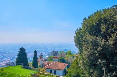 The roof and bell tower of San Vigilio Church against the hazy landscape from Castello di San Vigilio Park, Bergamo, Italy clipart