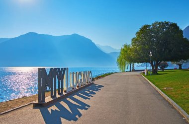 LUGANO, SWitzERLAND - 22 Mart 2022: Lugano Gölü kıyısı MyLugano yerleşim alanı su yüzeyinde güneş yolu önünde ve arka planda puslu Monte Sighignola, Lugano, Ticino, İsviçre