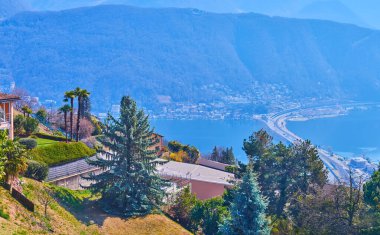 Carona 'nın güzel yeşil bahçeleri Lugano Gölü önünde uzun kozalaklı ağaçlar Melide Causeway ve Monte Sighignola yamacı, Ticino, İsviçre