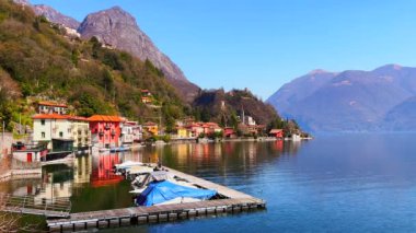 Lugano Gölü kıyısında demirli tekneleri olan küçük tersane San Mamete, Valsolda, İtalya 'daki dağlara ve renkli evlere karşı.