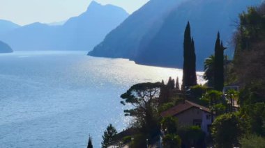 Lugano Gölü 'nde gün batımının tadını çıkarın. Puslu Alp siluetleri ve ön planda çam ve selvi ağaçları olan Oria bahçesi, Valsolda, İtalya.