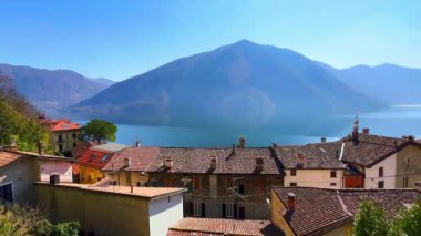 Lugano Gölü 'nün parlak mavi yüzeyi ve Lugano Prealps, Valsolda, İtalya' nın puslu Monte Pizzerno 'suna karşı Ortaçağ fayans çatıları ve evler Panorama
