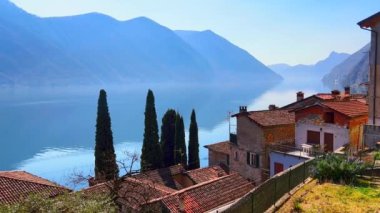 Lugano Gölü kıyısında tarihi evleri olan küçük bir Oria köyünün panoraması. Selvi ve puslu dağların arasında San Sebastiano Kilisesi, Valsolda, İtalya