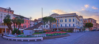 DESENZANO DEL GARDA, ITALY - APRIL 10, 2022: The evening Piazza Giacomo Matteotti with monument to Brave Aviators, surrounded with tulip flower beds, historic hotels and restaurants, on April 10 in Desenzano del Garda