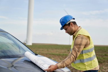 Rüzgar türbini sahasında duran Latin erkek mühendis ve araba kaputu üzerindeki kağıt projesini kontrol ediyor