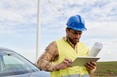 Latin erkek mühendis dijital tablet tutuyor ve kağıt projesiyle rüzgar türbini sahasında duruyor. 
