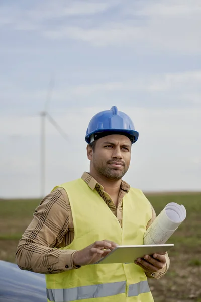 持有数码平板电脑的拉丁男性工程师 他们站在风力涡轮机领域的纸张项目上 — 图库照片