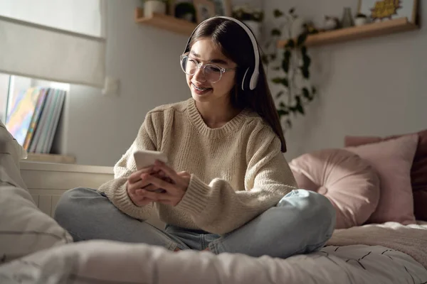 Caucasian teenage girl browsing phone with smile and wearing headphones while sitting on bed