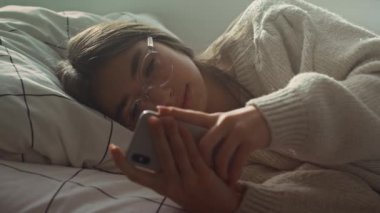 Kafkasyalı genç kızın yatakta yan yatarken telefonuna göz gezdirirken yakın çekim. 8K 'da kırmızı helyum kamerayla çekildi..  