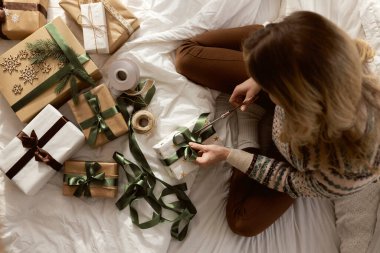Beyaz kadın yatakta oturuyor ve evde Noel hediyesi paketliyor. 