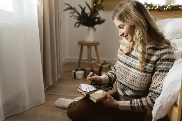 朗らかな白人女性が床に座ってプレゼントの中にクリスマスカードを書いて — ストック写真