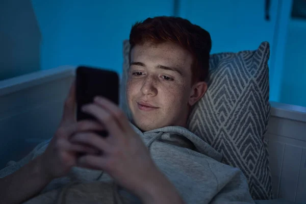 Vidám Kaukázusi Tinédzser Fiú Mobiltelefont Használ Miközben Éjjel Ágyban Fekszik — Stock Fotó