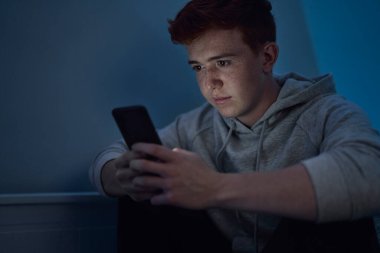 Geceleri odasında otururken cep telefonu kullanan beyaz bir çocuğa odaklan. 