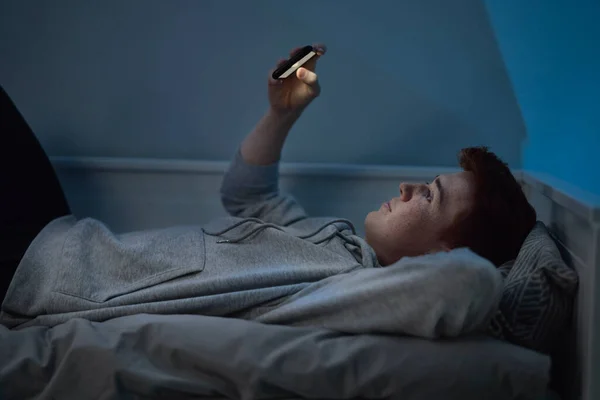 快乐的高加索少年晚上在房间里躺在床上看手机 — 图库照片