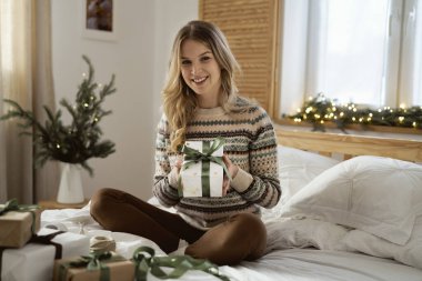 Yatakta otururken elinde Noel hediyesi tutan neşeli beyaz kadın portresi.