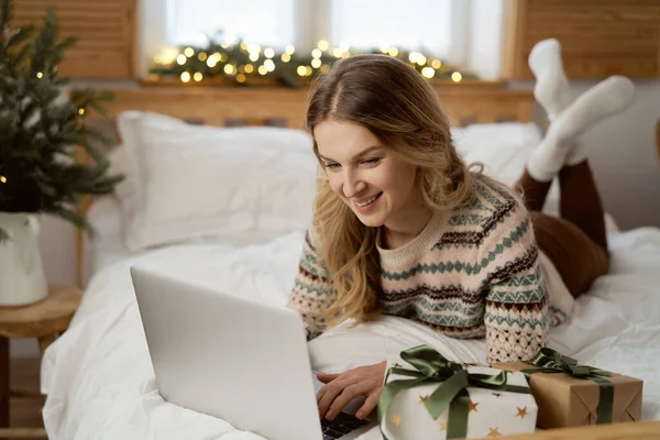 Кавказская Женщина Лежит Постели Просматривает Ноутбук Среди Рождественских Подарков — стоковое фото